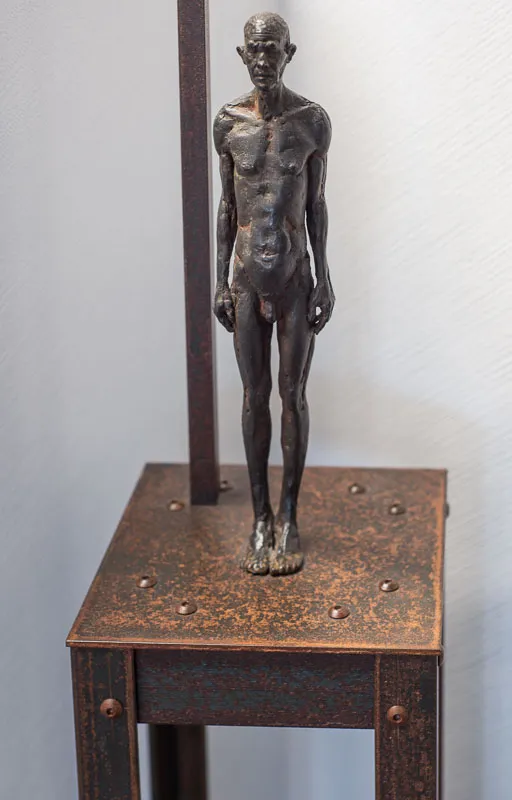 Bronze male figure on a narrow but tall table, beside a flag pole.