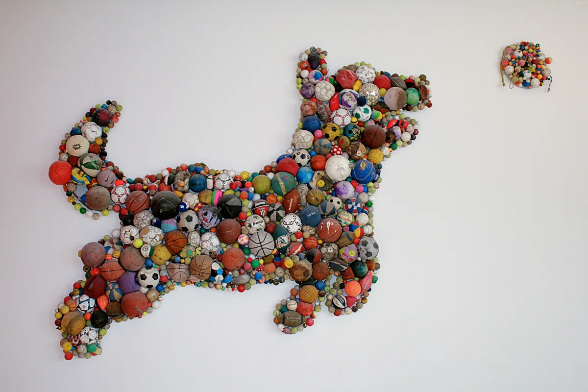 A bas relief sculpture of a dog chasing a ball, made out of weathered balls of various kinds.