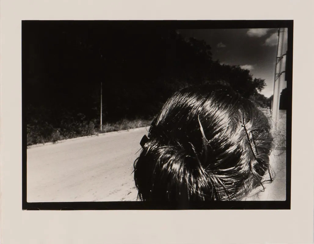 Black and white photograph o the back of a woman's head; a street scene in the background.