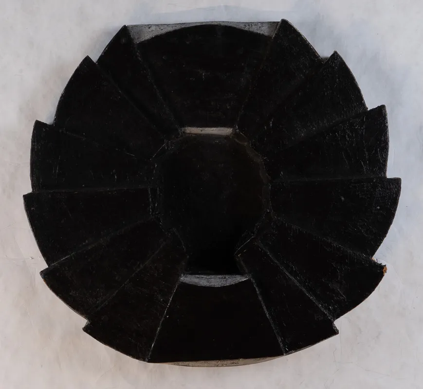 Black disc in wood with what apepars to be stylized laurel leaf decoration around outside.