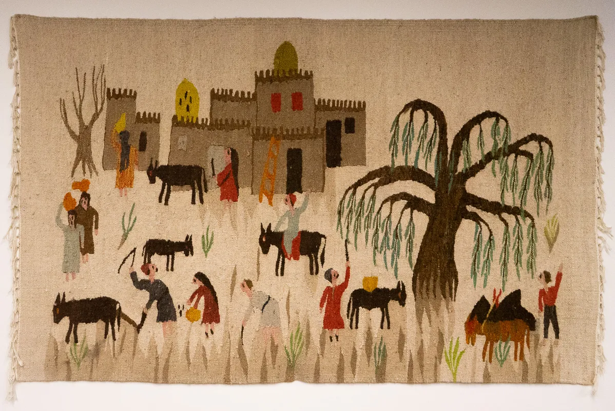 Tapestry/carpet of a scene of people harvesting crops under a large willow tree. A building in the background.